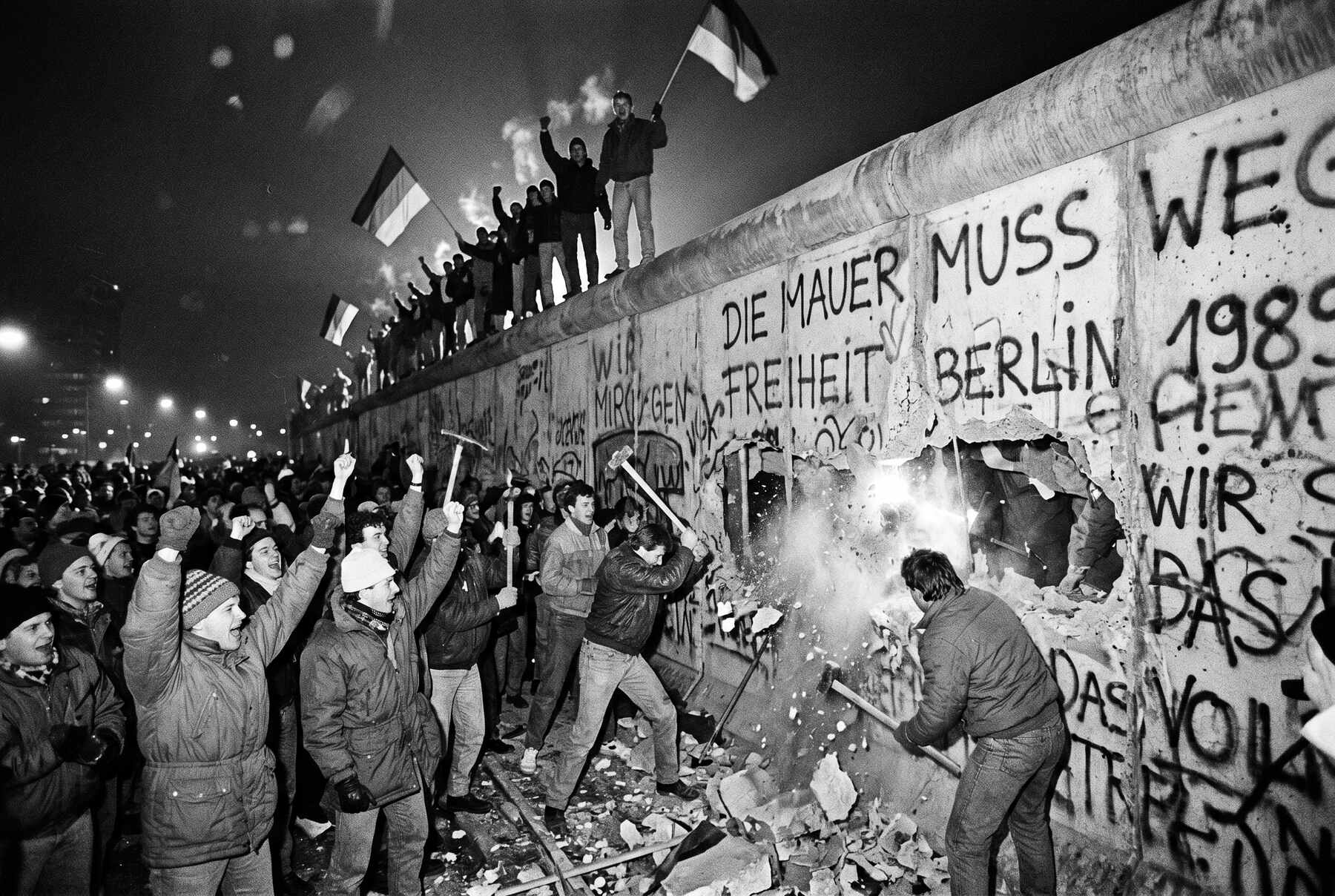 Crowds at the Berlin Wall as it comes down in 1989, figures climbing and passing through the breached concrete.